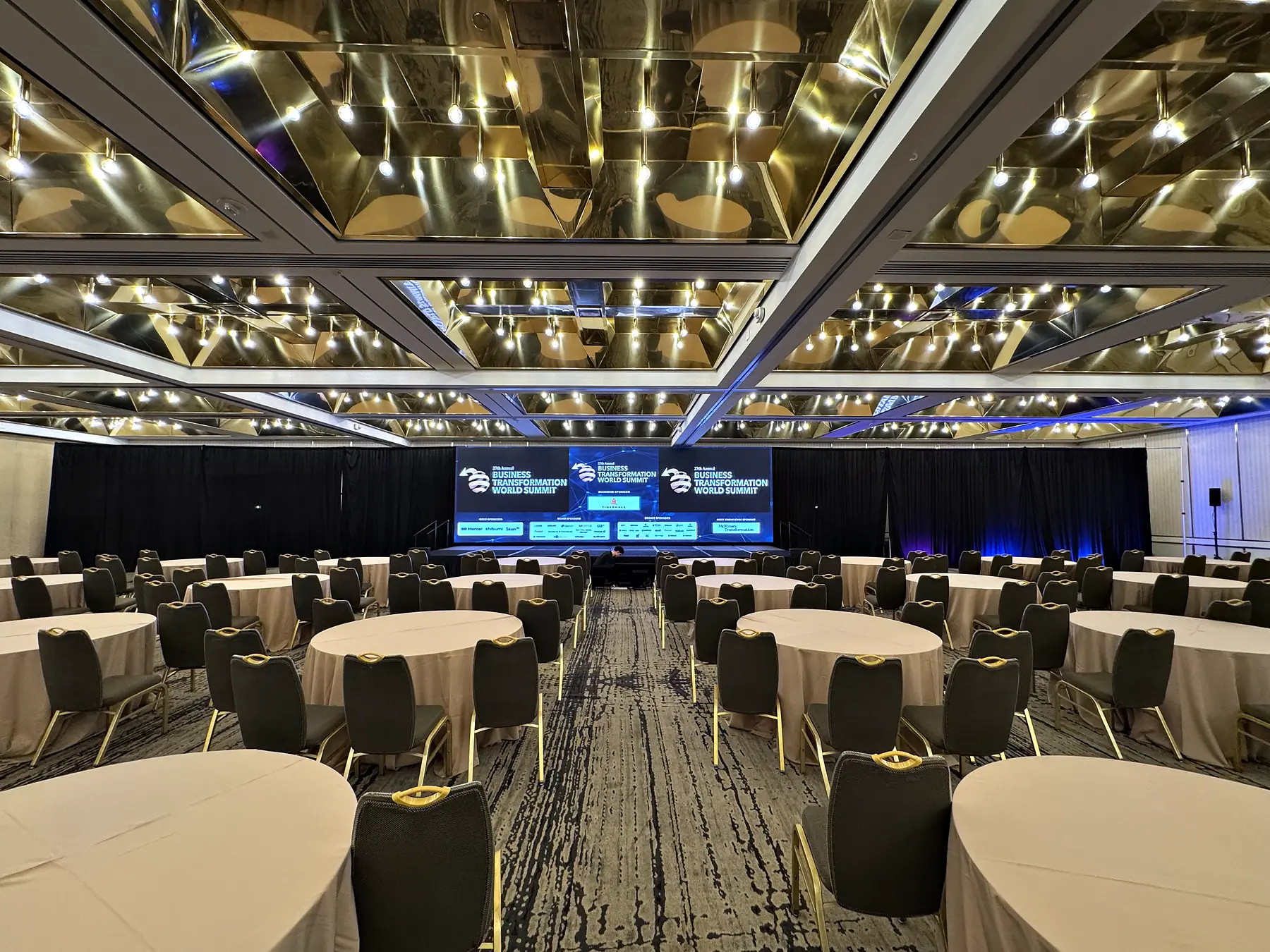 A ballroom set with round tables under a coffered gold ceiling of recessed lights, black drape along the far wall, and a wide video wall on a low stage at the end of the room carrying a title graphic.