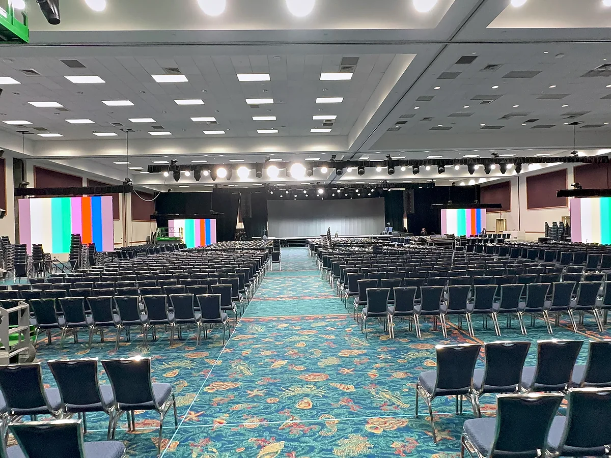 A large room set in rows of chairs facing a low stage, a wide video wall standing at each side of it carrying colour bars, a lighting rig hung across the front and a scissor lift at the edge of the frame.