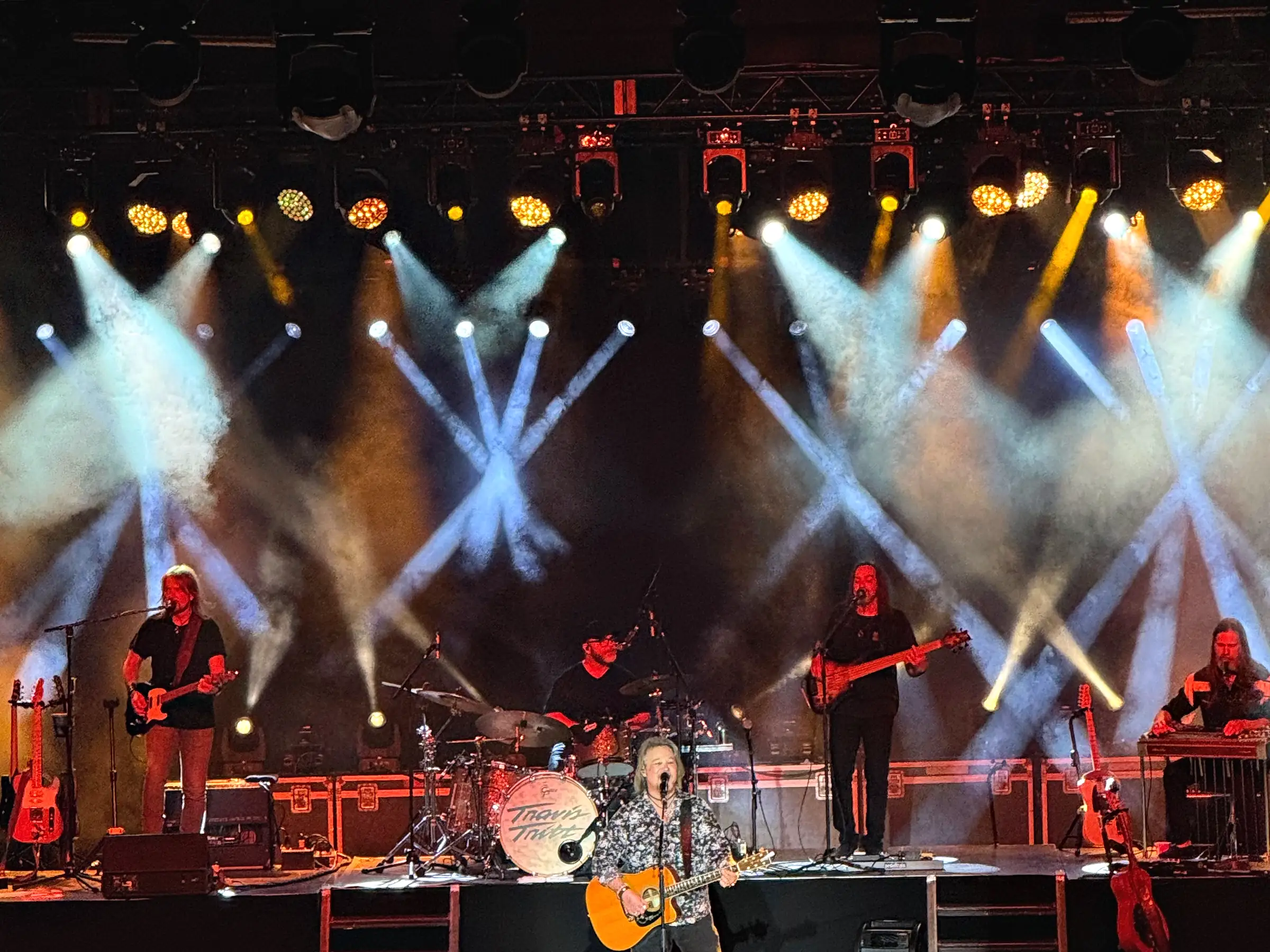 A band playing across a covered stage under a dense rig of moving lights throwing crossed beams forward through haze, silhouetted heads of the audience along the bottom of the frame.