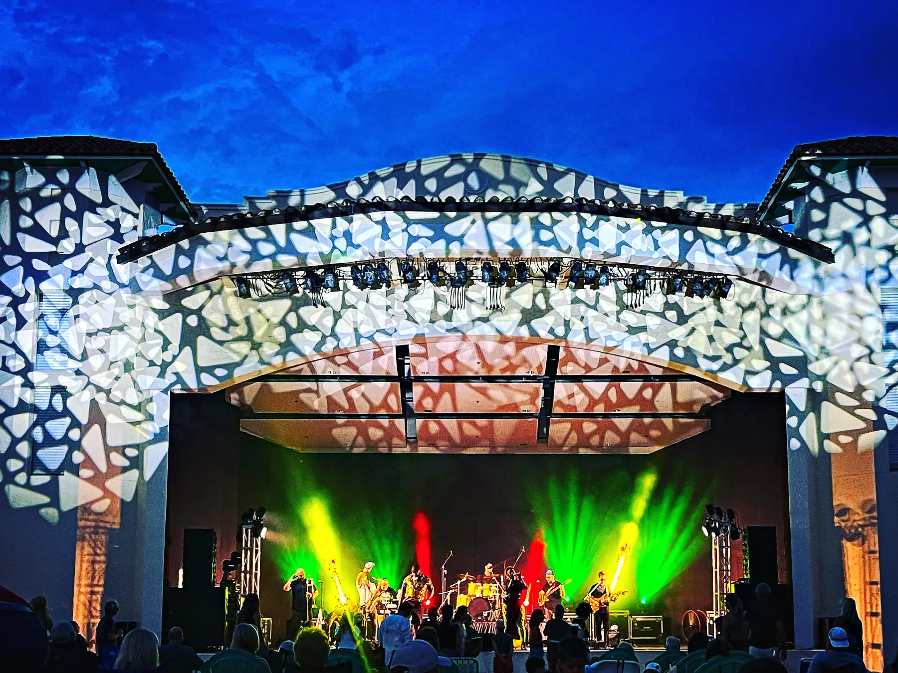 An open-air bandshell at dusk with a broken pattern of light thrown across its whole facade and roof, a band playing under green and red beams on the stage inside it, and the silhouettes of an audience seated on the grass in front.