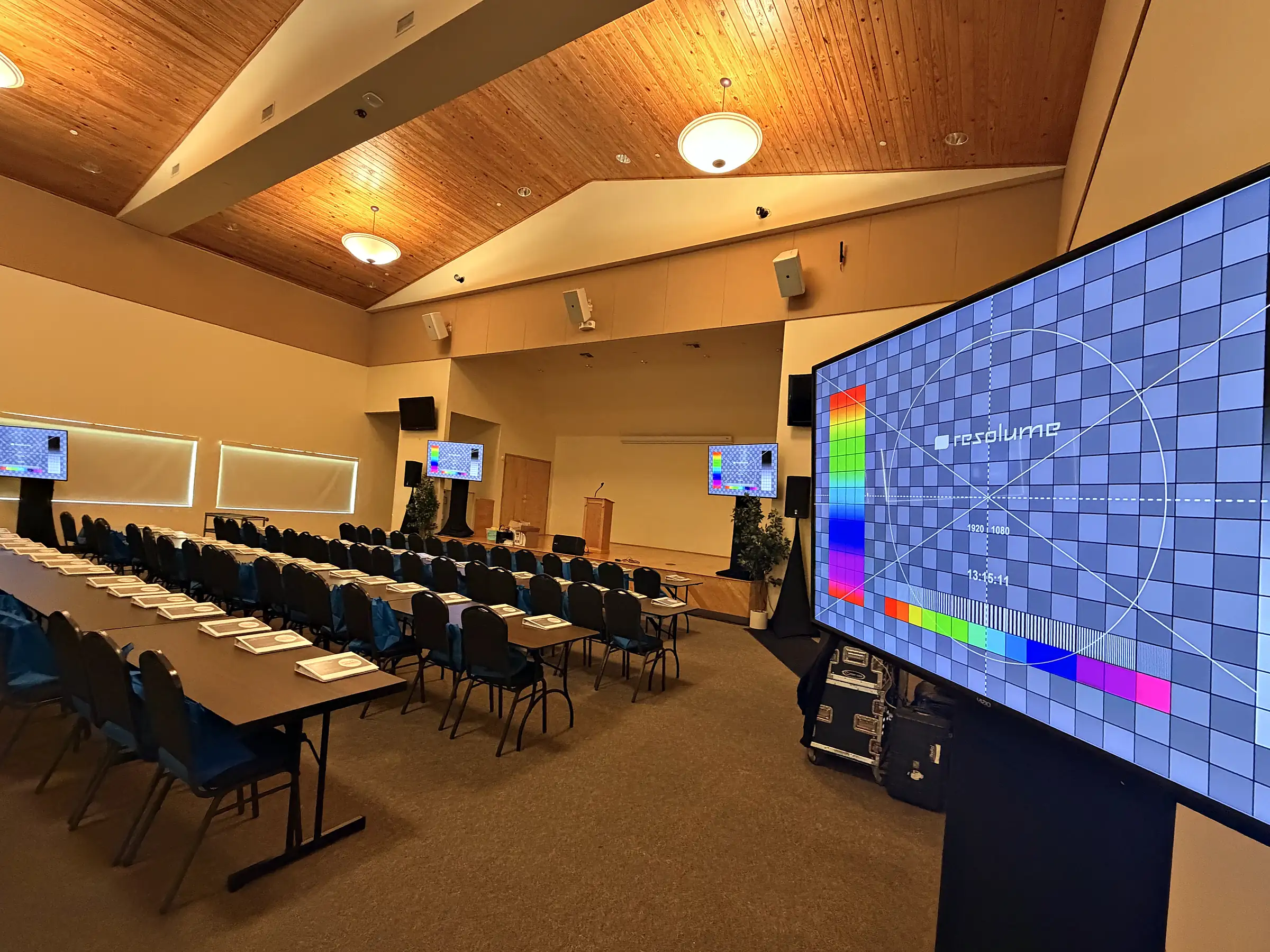 A breakout room under a pitched wooden ceiling, long tables set with pads and chairs facing a large screen carrying a grid test pattern, loudspeakers on stands at both sides and road cases still on the floor.