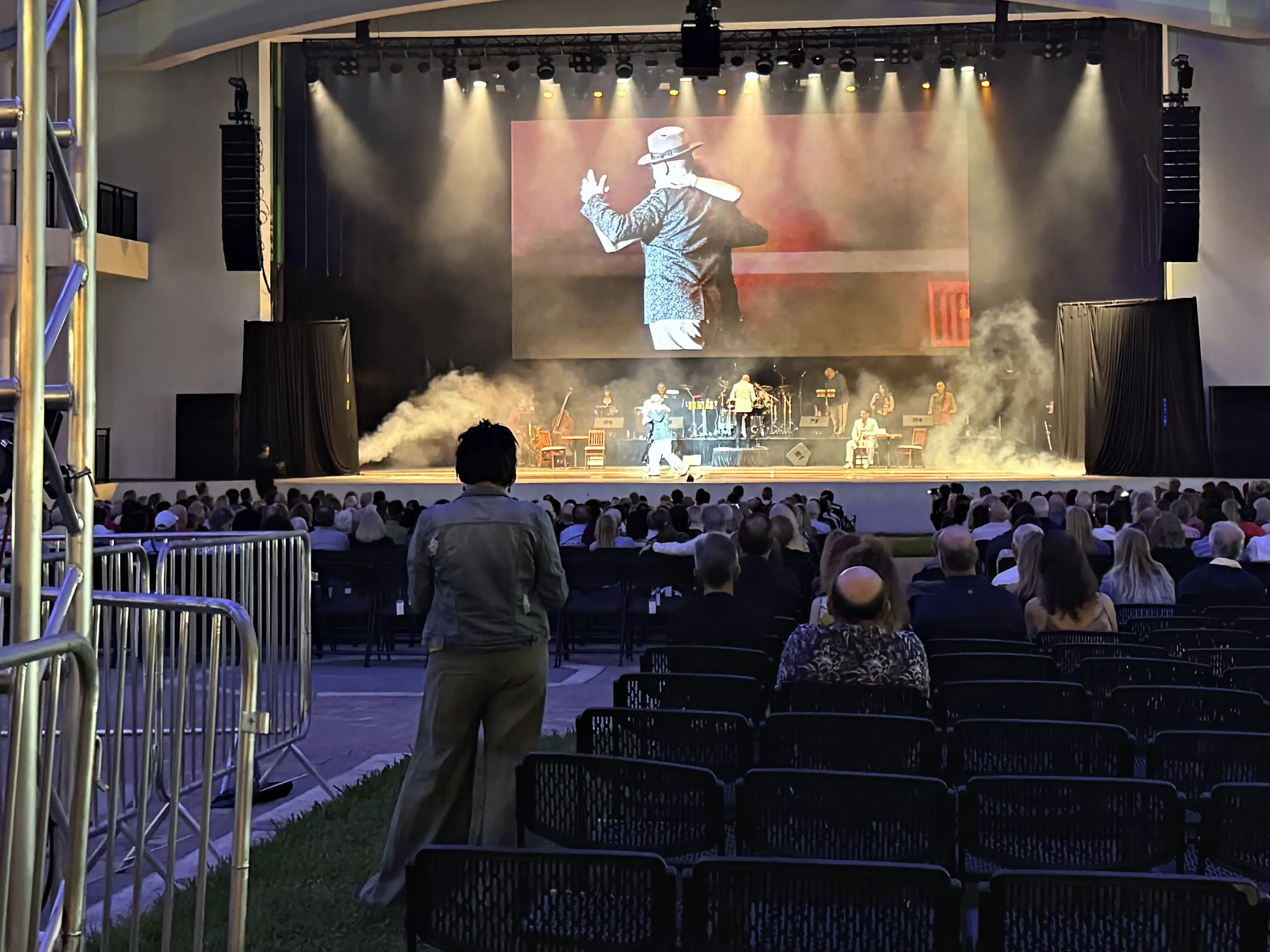 A stage under a canopy roof with a wide video wall above it carrying a close shot of the performer, beams of light crossing through haze, musicians and their instruments across the back of the deck, and a seated audience filling the rows in front.
