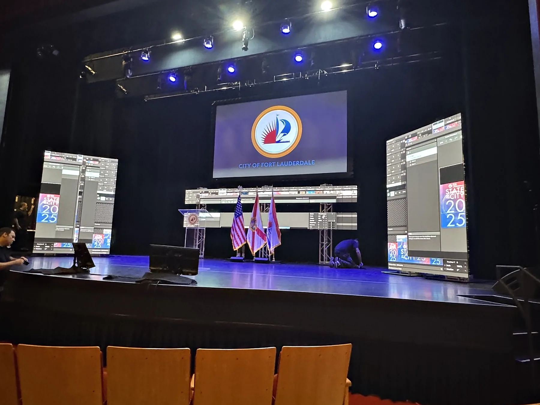 A theater stage set for a civic address: a lectern at the left, three flags on stands at the center, a wide screen above the stage carrying a municipal seal, and tall portrait screens standing at either side.