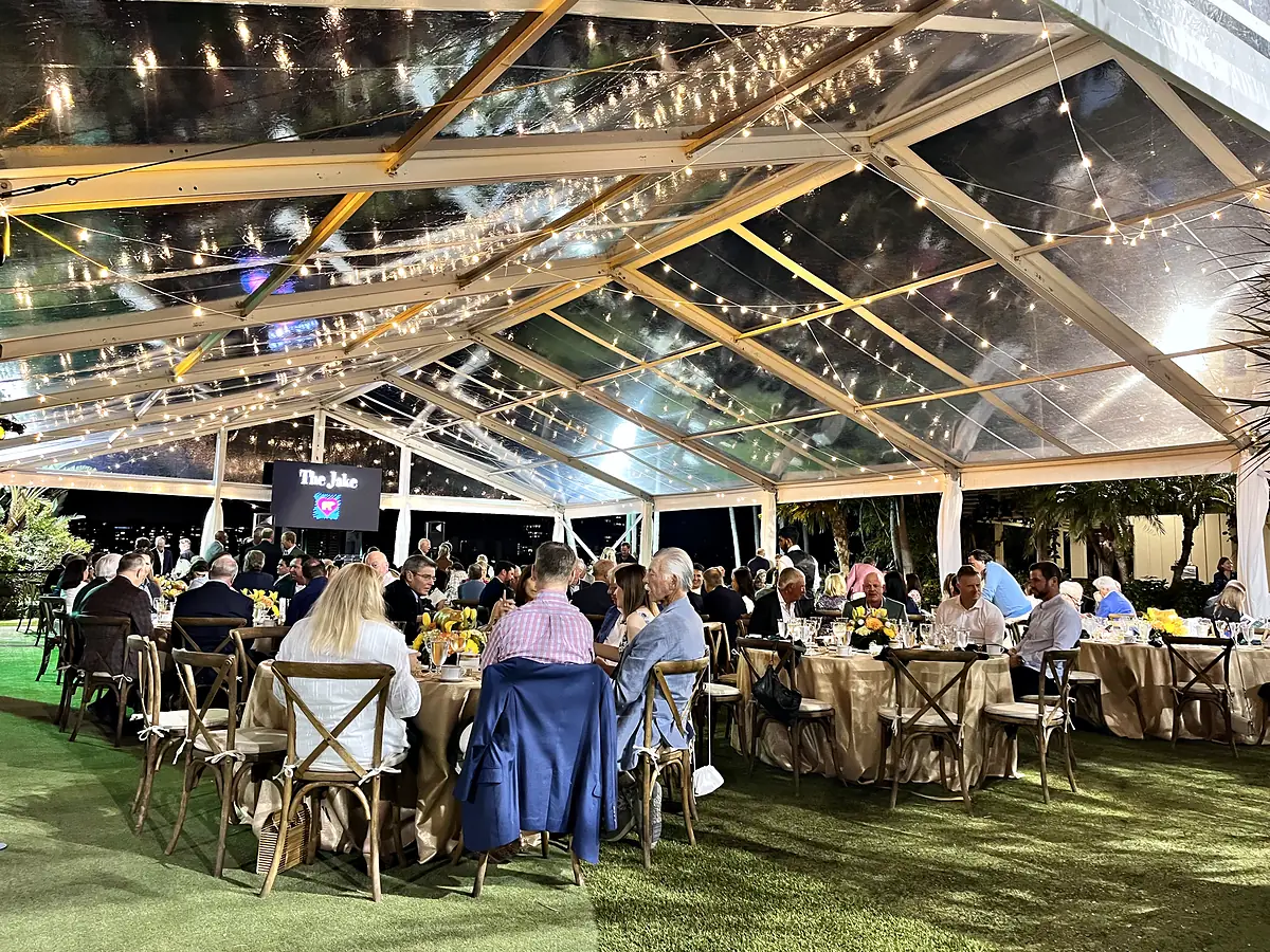 A dinner laid on round tables under a clear-topped tent on grass at night, strings of small lights running the length of the ceiling, and a screen on a stand at the far end of the room.