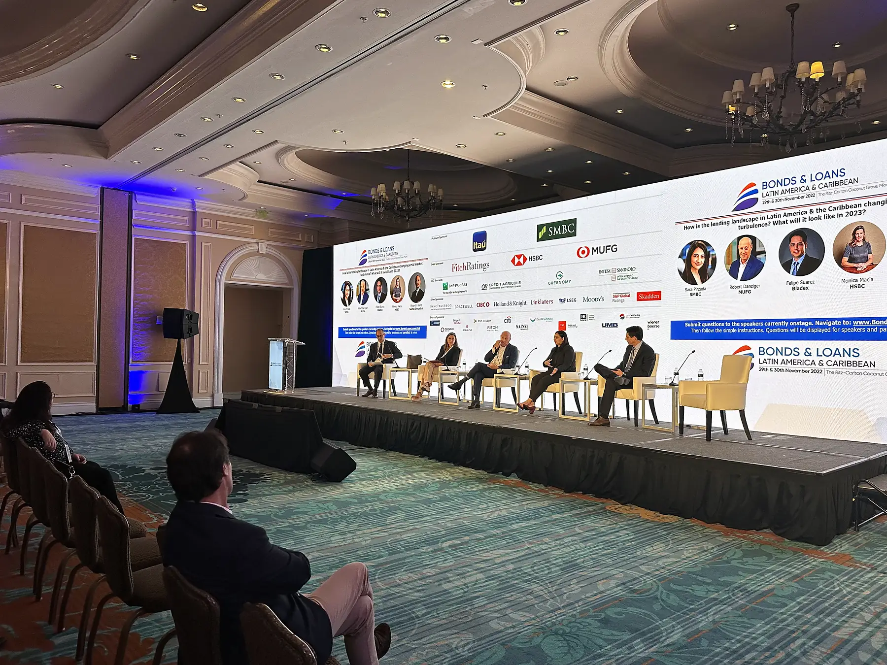 A panel seated in armchairs along a low stage running the width of a chandeliered ballroom, a wide screen behind them carrying a row of sponsor marks, uplight on the walls and an audience seated at rounds and in rows in front of it.