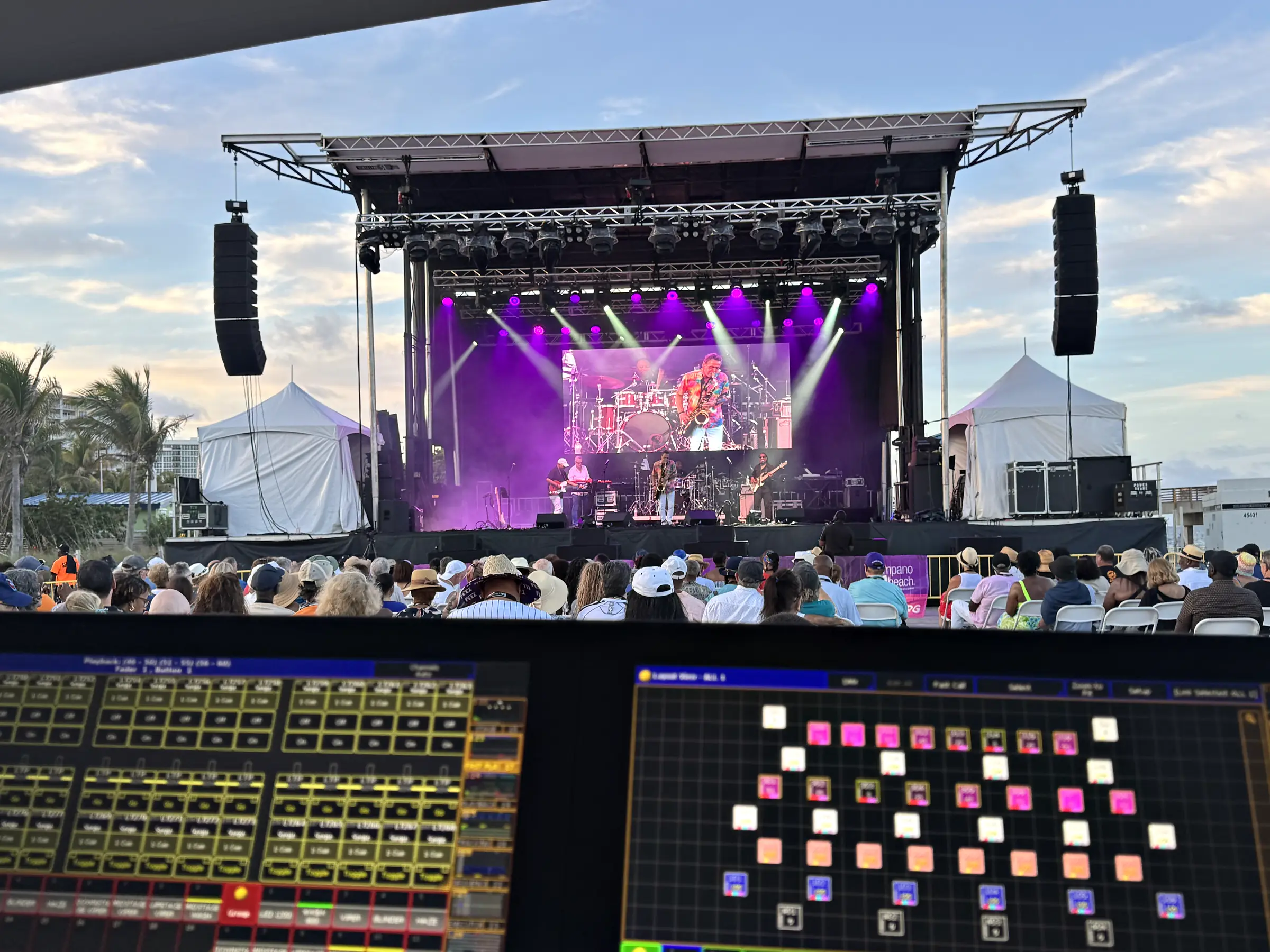 Two lit control surfaces across the bottom of the frame, and beyond them an outdoor stage under a trussed roof with loudspeakers flown at both sides, a band playing under coloured beams and a seated audience between the desks and the deck.