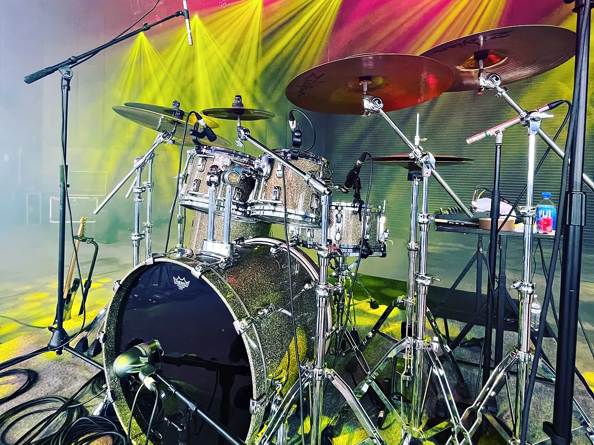 A drum kit set up at the center of a stage with microphones on short stands around it and cymbals catching the light, magenta and yellow beams crossing the backdrop behind it and cable run across the deck in front.