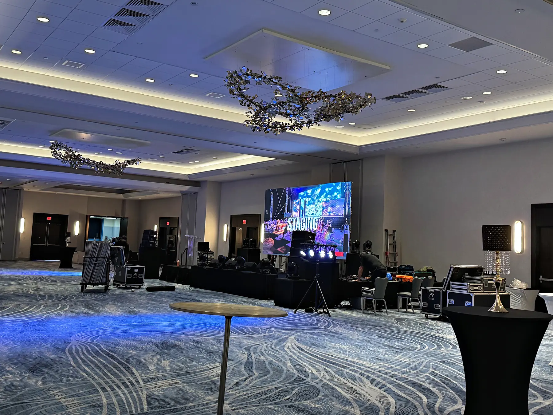 An empty hotel ballroom lit blue, a small LED screen reading “Staging” beside a bank of moving lights and stacked loudspeaker cabinets, decorative fixtures hanging from the ceiling and road cases spread across the carpet.