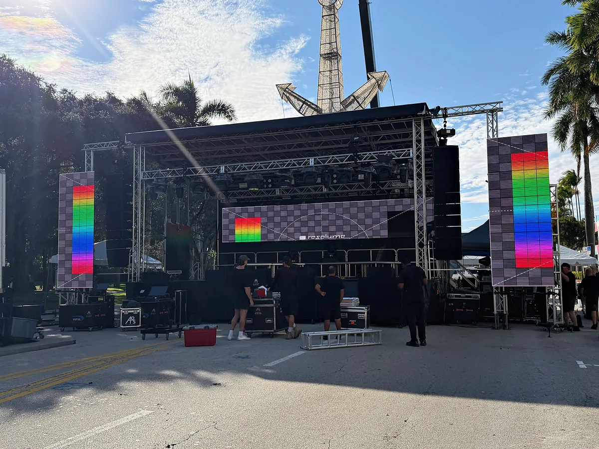 An outdoor stage under a mobile roof with a video wall across the back and a tall portrait video tower standing at each side, all three carrying color test fields, palms above and crew and road cases working around the base in daylight.