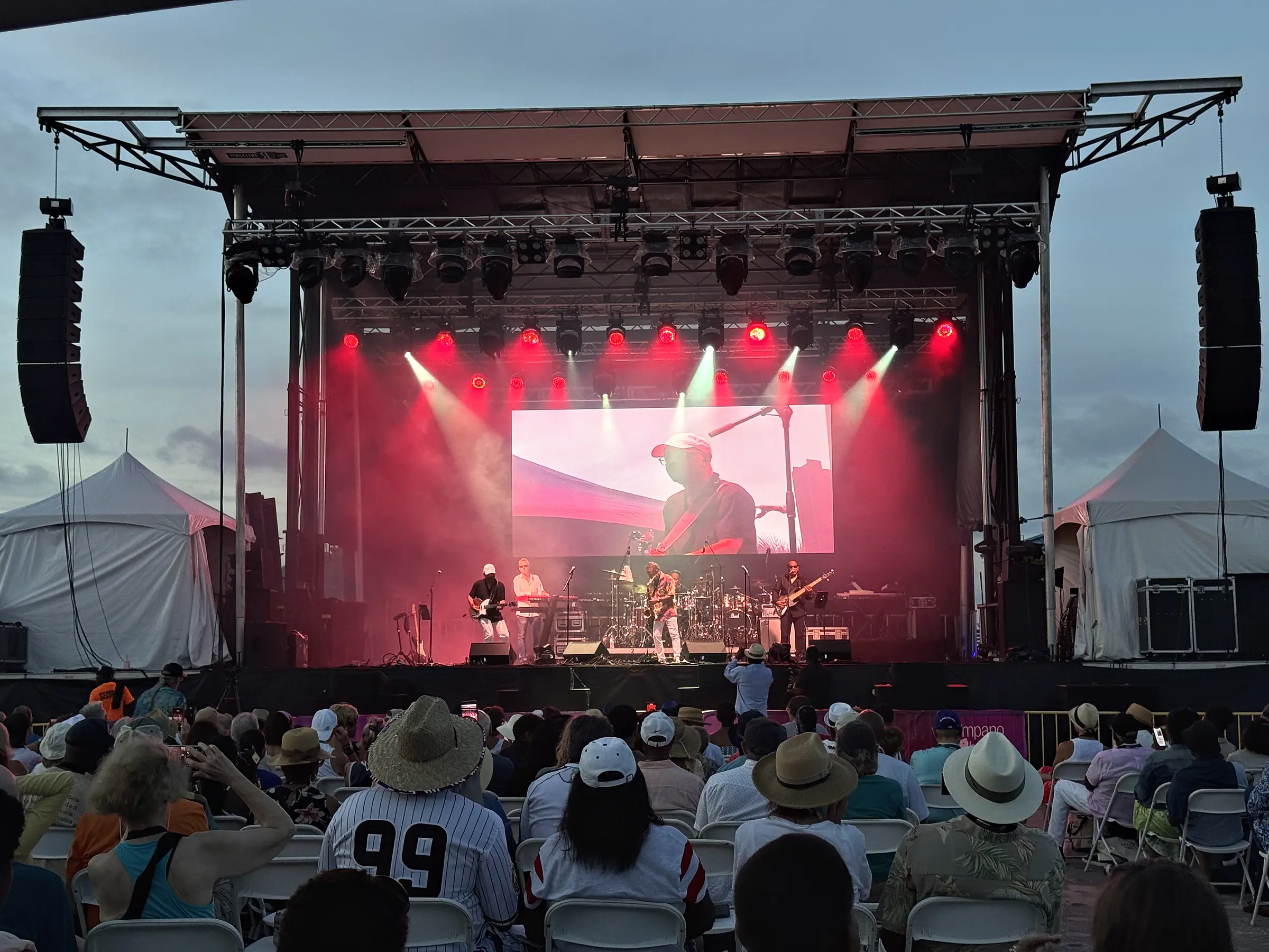 An outdoor stage under a trussed roof lit red and white, loudspeakers flown at both sides of it, a band across the deck with a wide screen behind them, and a crowd filling the ground in front in hats and caps.