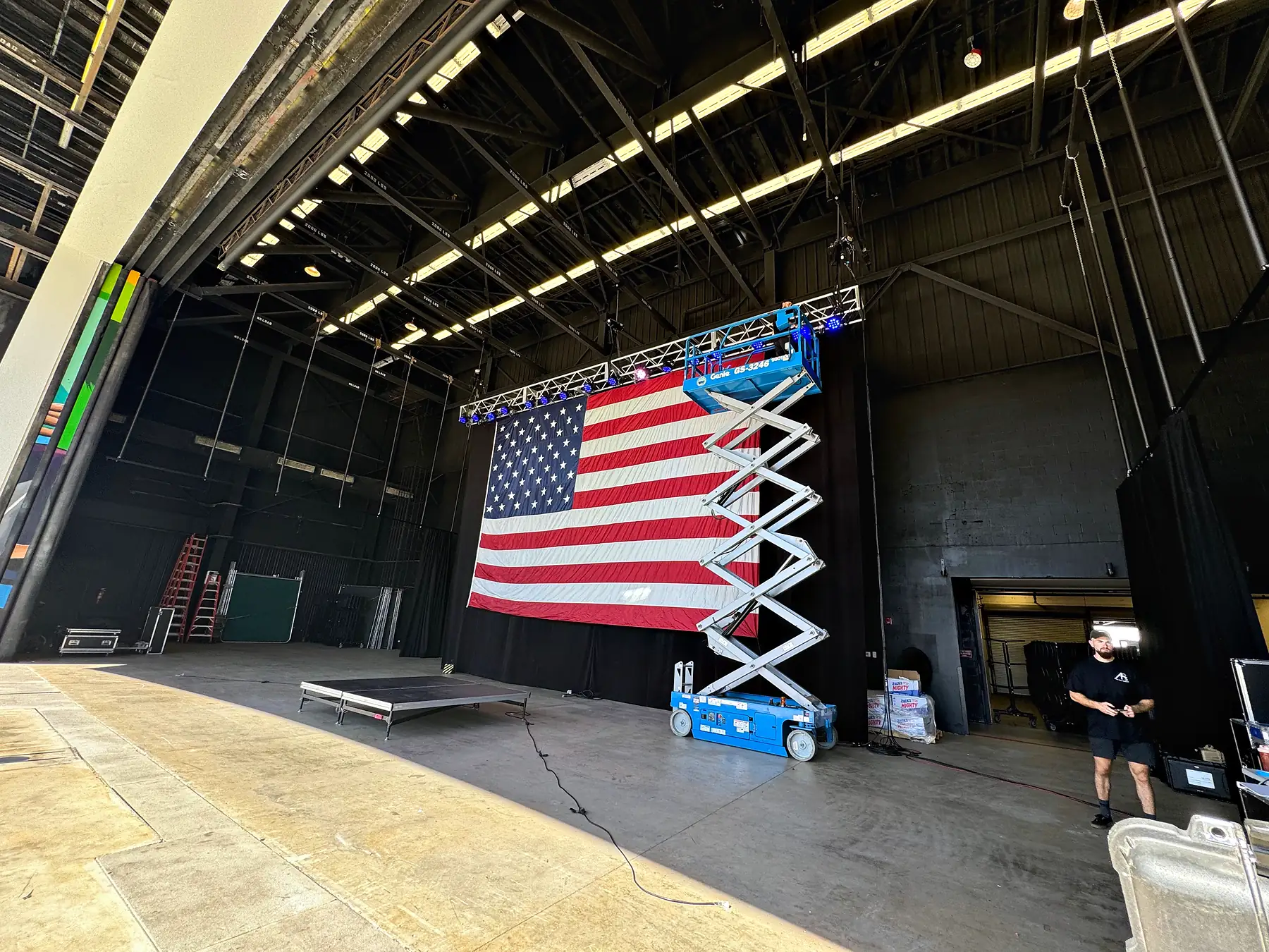 A large flag hung across the back wall of a covered stage with a scissor lift standing in front of it, steel roof structure overhead, a crew member working at the stage edge and the deck otherwise empty.