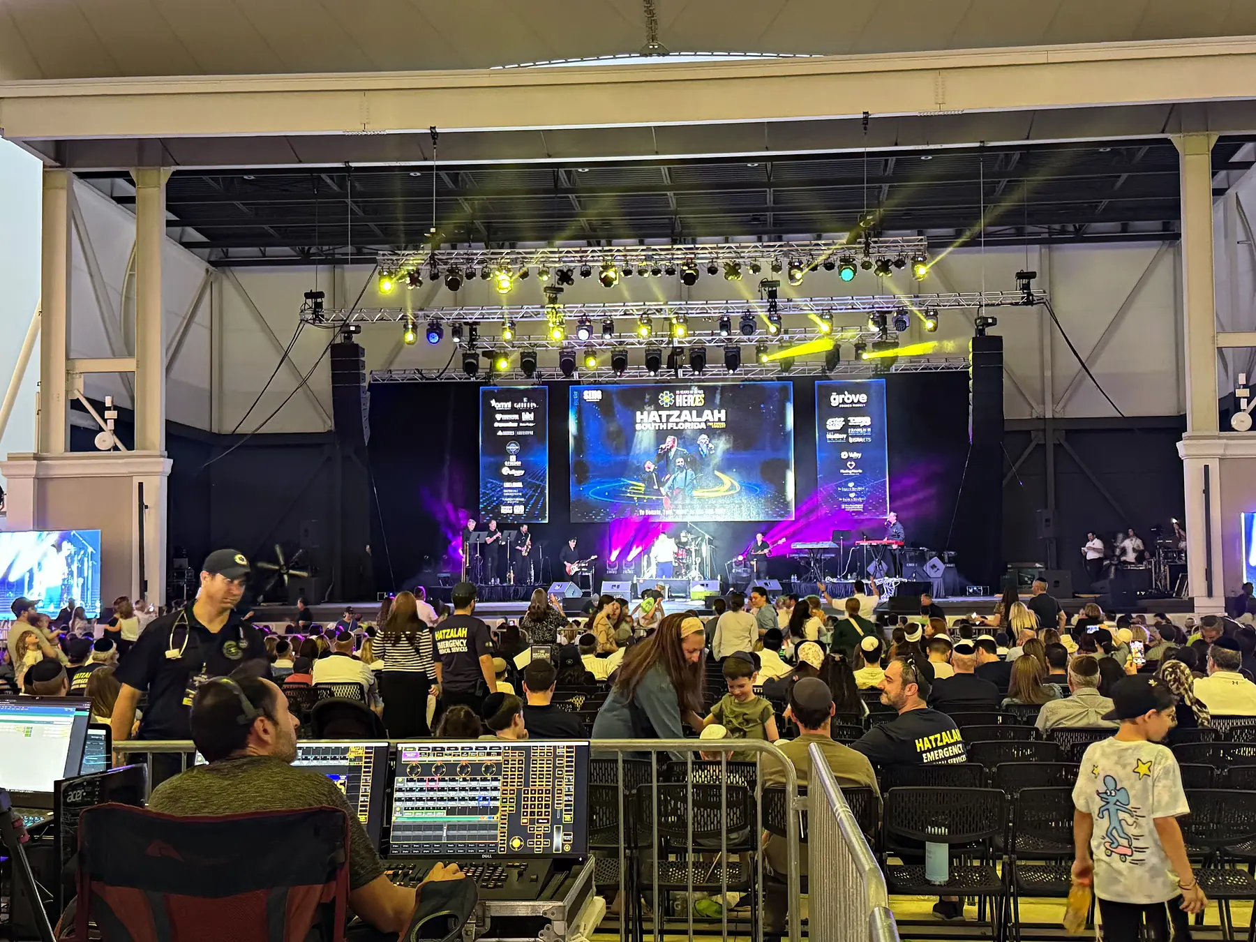 A front-of-house position under a covered pavilion, a lighting console and an audio console lit in the foreground, a standing audience beyond them and a band on a stage under moving lights at the far end of the room.