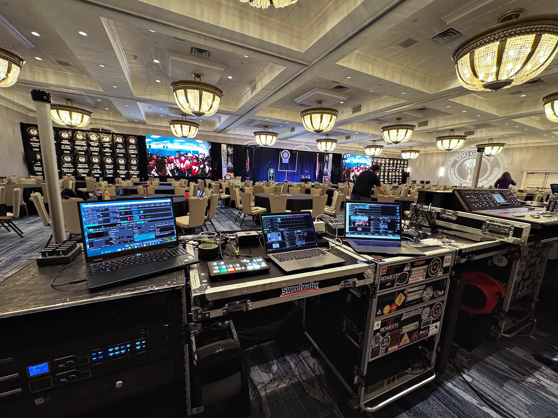 A front-of-house position built out of road cases, three laptops and a console surface lit under a coffered ceiling, with the room beyond it carrying video walls and rows of guests at tables.