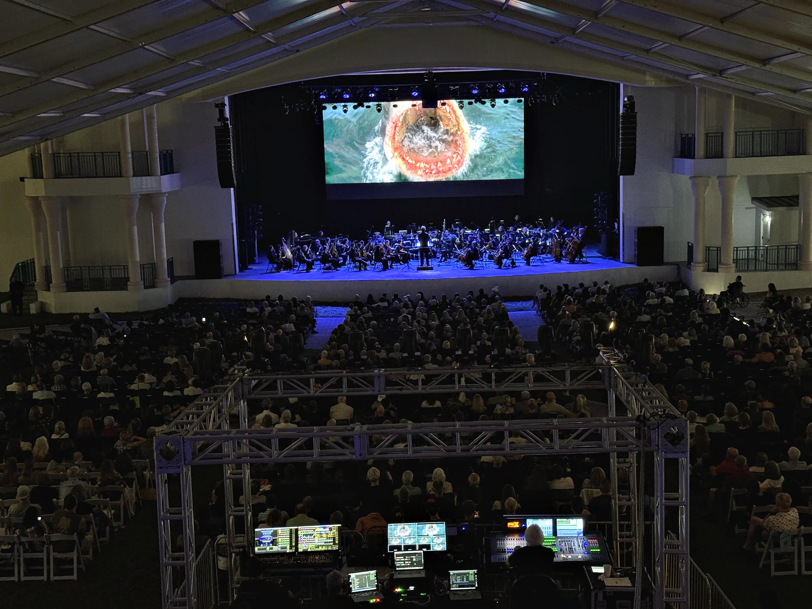 An audience filling every row under a canopy roof, seen from behind a front-of-house position built inside a cage of truss with lit control surfaces across it, and beyond them an orchestra on a lit stage below a large screen.
