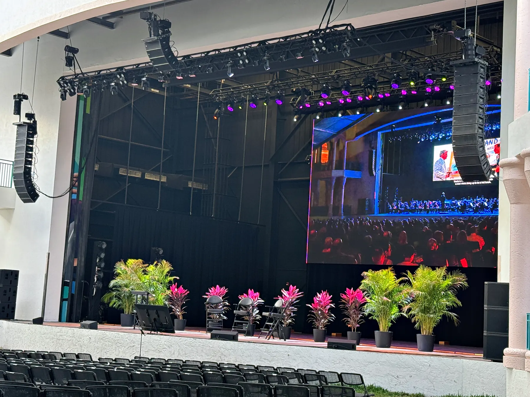 A covered outdoor stage with a line array flown at each side, a video wall upstage carrying an image, planted flowers in a row along the stage edge and rows of empty seats in front of it.