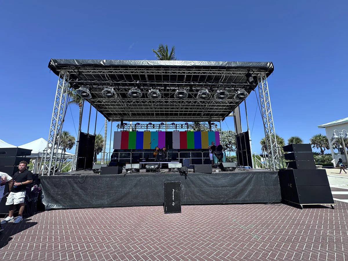 A mobile stage standing on a brick plaza under a clear sky, its roof carried on truss towers with lighting fixtures hung across it, loudspeakers stacked at both sides of the deck and a video wall on colour bars at the back of it.