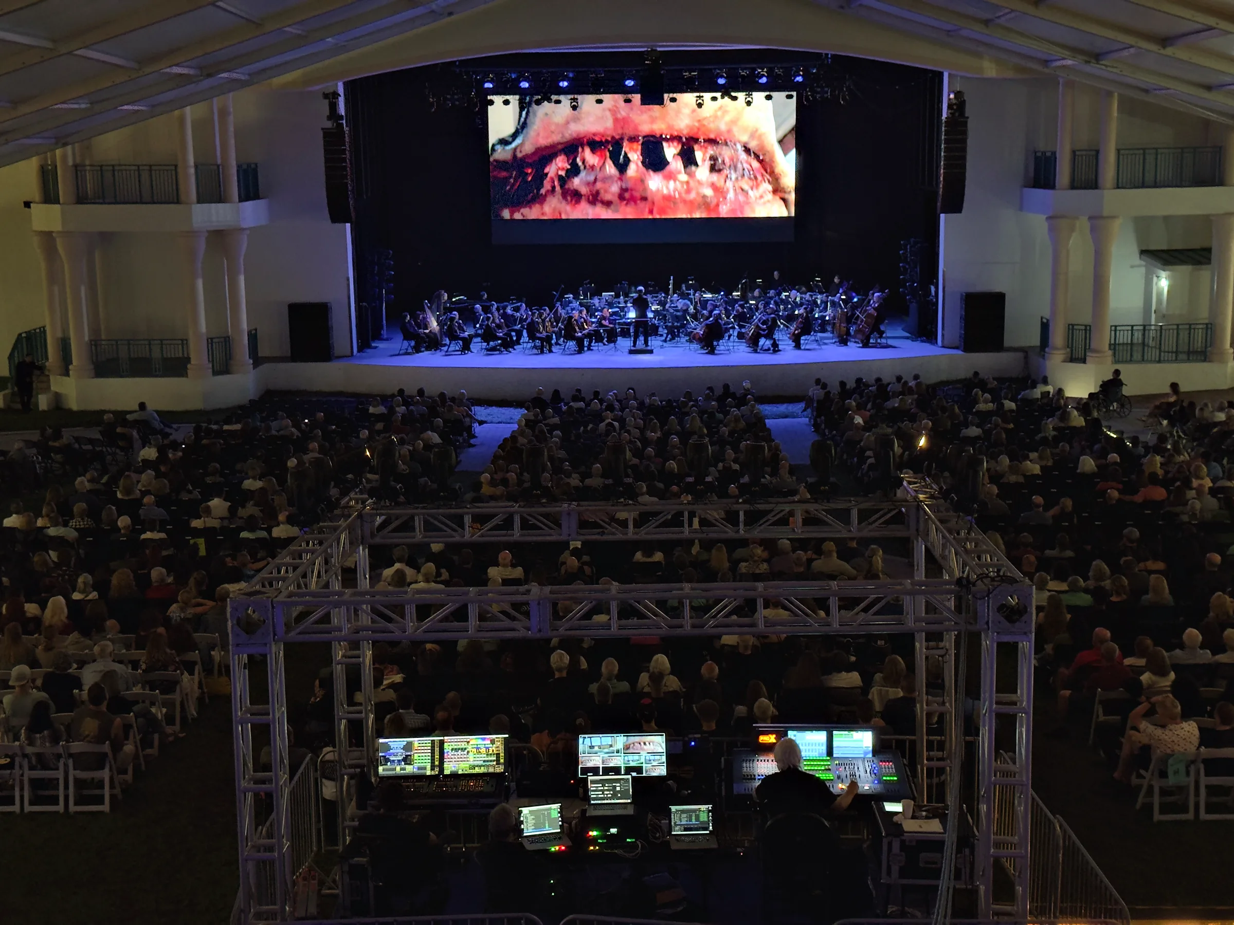 An orchestra playing on a stage below a large video wall carrying a film still, a full house seated in front of it, and a front-of-house position built inside a truss structure in the foreground.