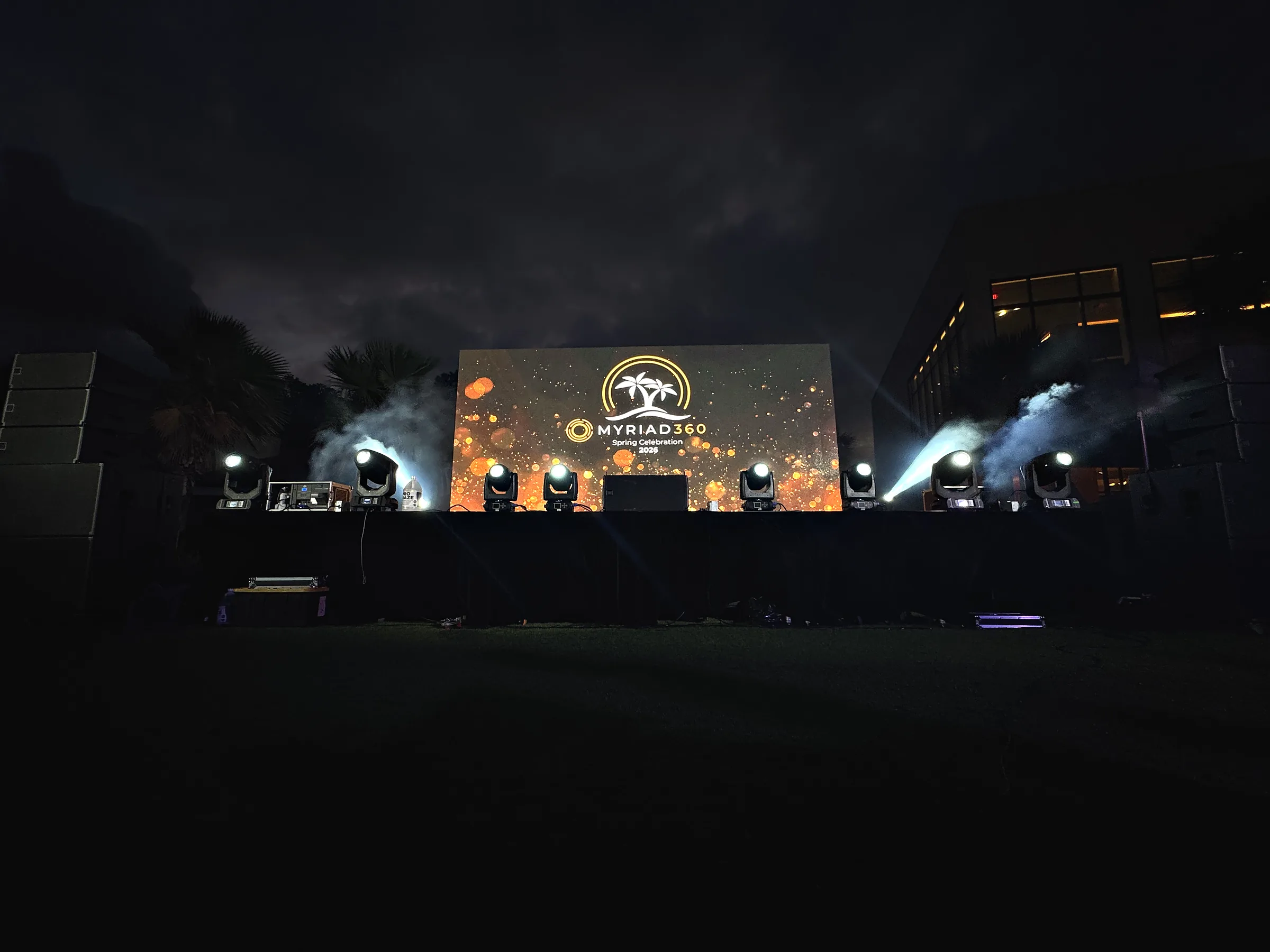 An outdoor stage at night carrying a wide video wall with a logo on it, beams of light cutting out through haze from fixtures along the front edge, and dark grass running away in the foreground.