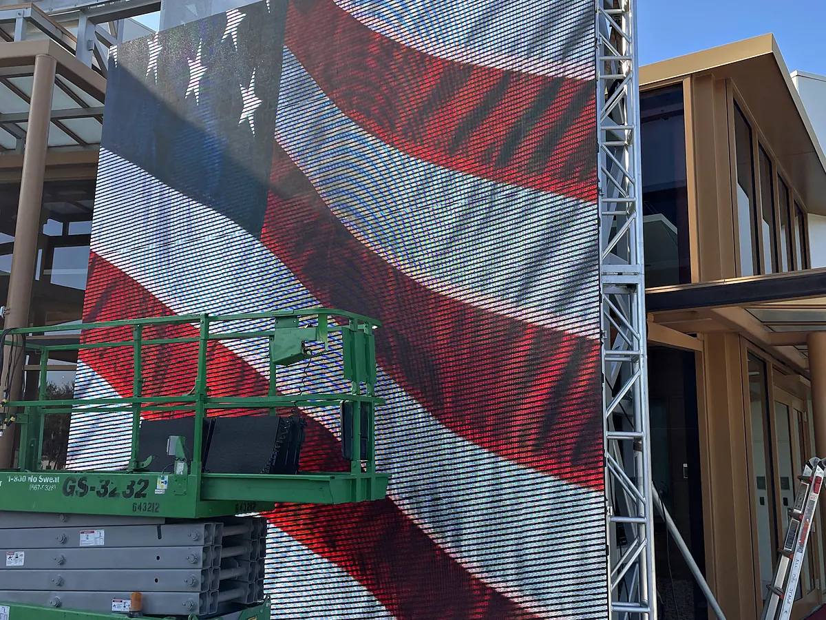 A tall video wall standing outdoors on ground-support truss carrying a flag image, a green scissor lift parked against the left of it, cable run out across the paving and a building with glass and timber behind.
