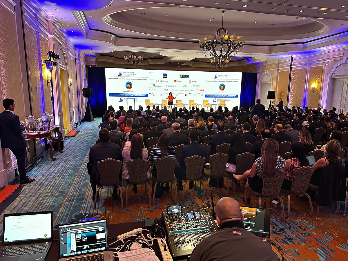 A full ballroom seen from behind the front-of-house position, an audio console and two laptops on a table in the foreground with an operator beside them, rows of seated people running away to a low stage where speakers sit in armchairs below a wide screen.