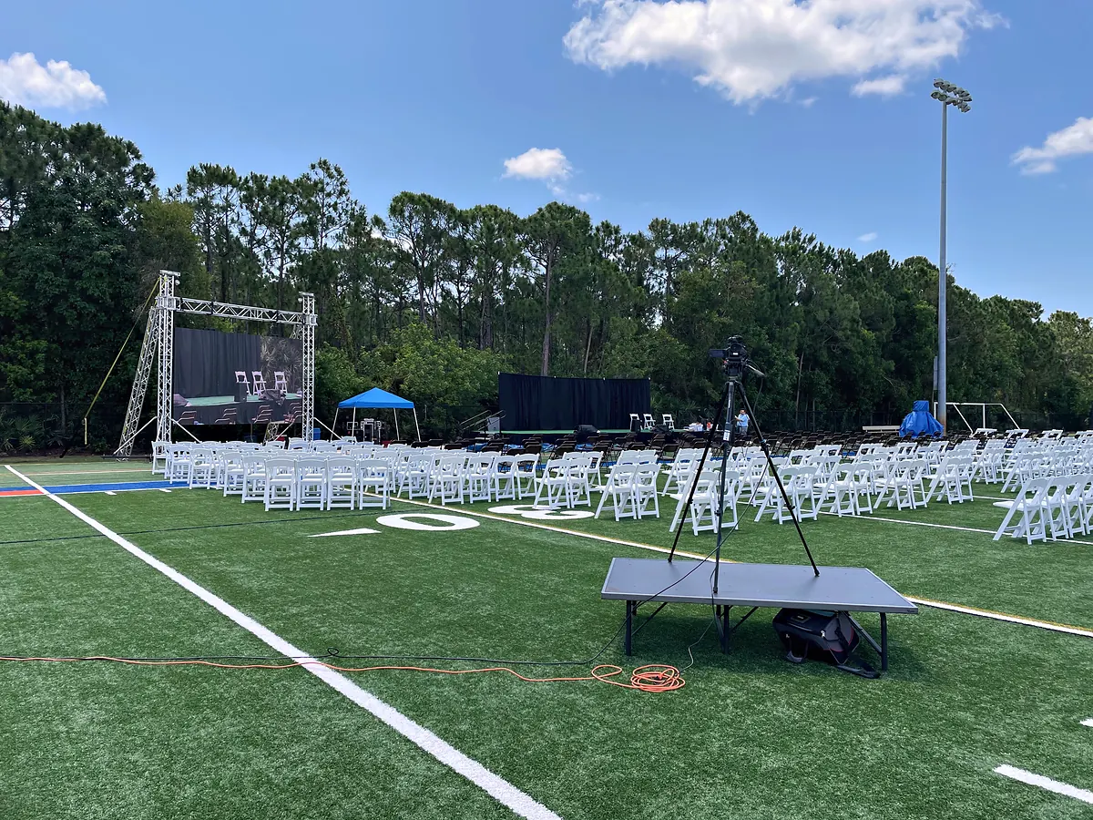 A covered stage on ground-supported truss built at one end of a marked sports field under a bright sky, rows of white folding chairs set out on the turf in front of it and a camera position on a platform at the side.