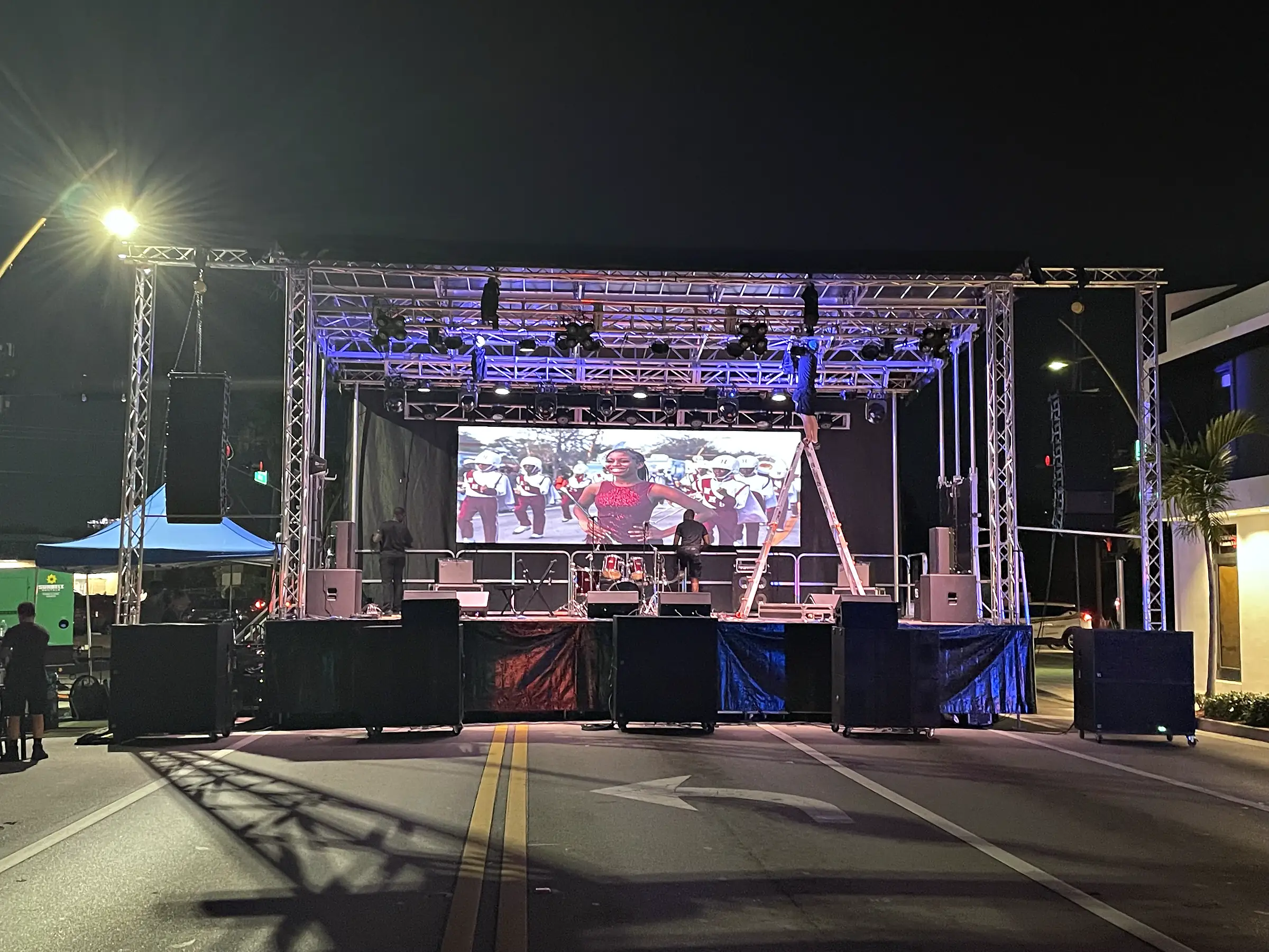A stage built on a closed street at night under a trussed roof, a video wall carrying an image across the back of it, subwoofers stacked along the front edge of the deck and lighting towers standing at either side.