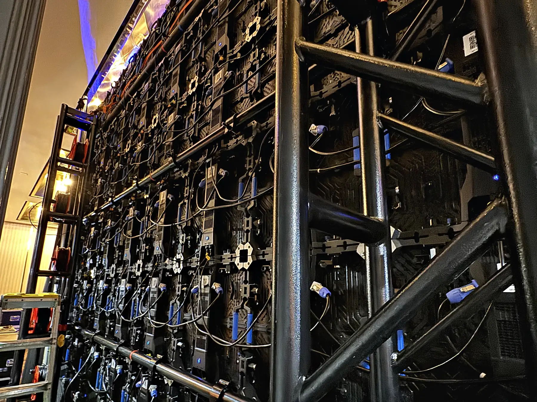 The back of a video wall seen from behind the ground-support truss carrying it, rows of panel frames with data and power cable looped between them, and a ladder standing at the side.