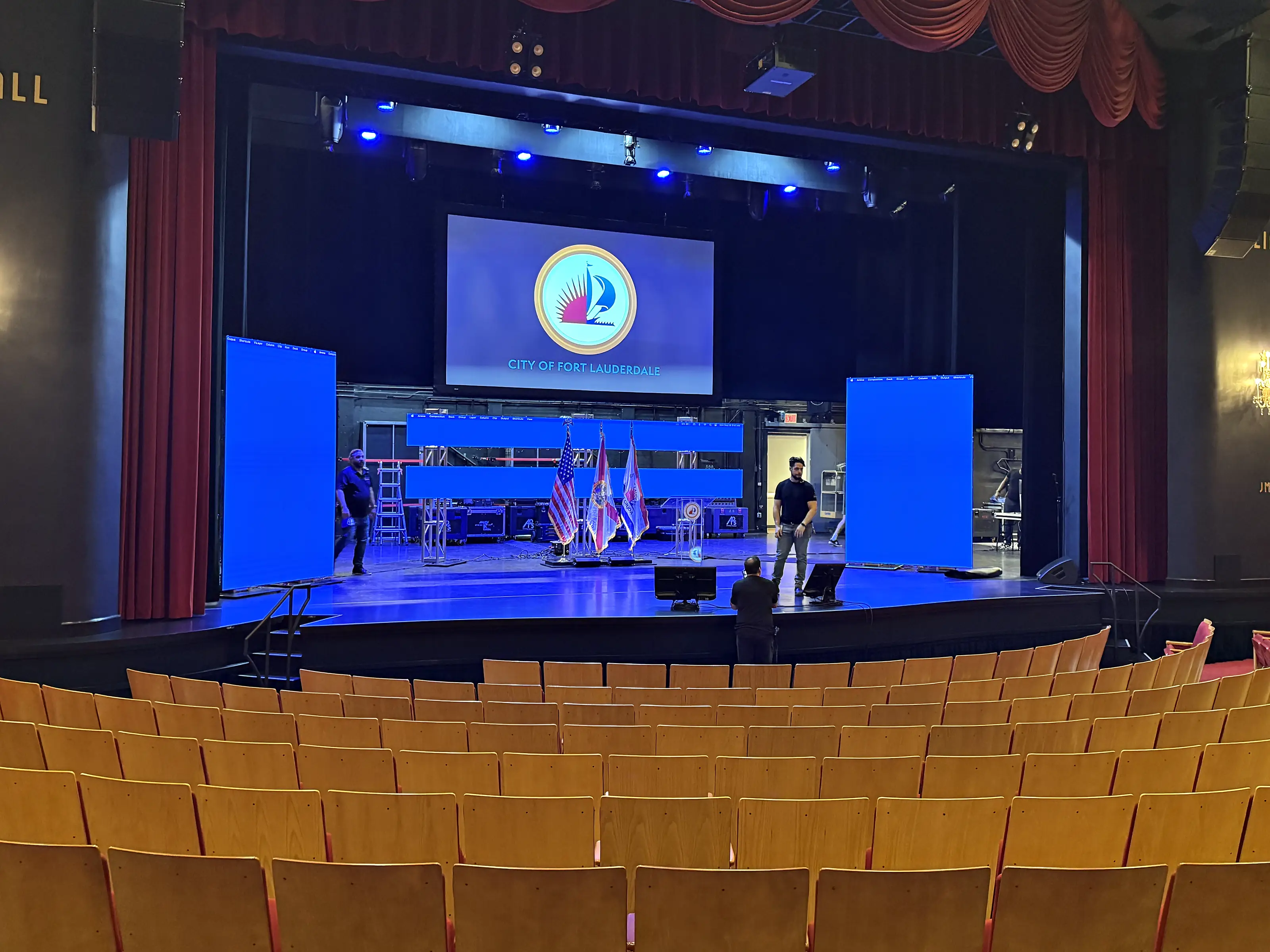 A theater stage seen from the back of the house, a wide screen hung at the centre of it and a portrait screen standing at each side, flags and a lectern on the deck, crew working at the back and rows of empty wooden seats in the foreground.