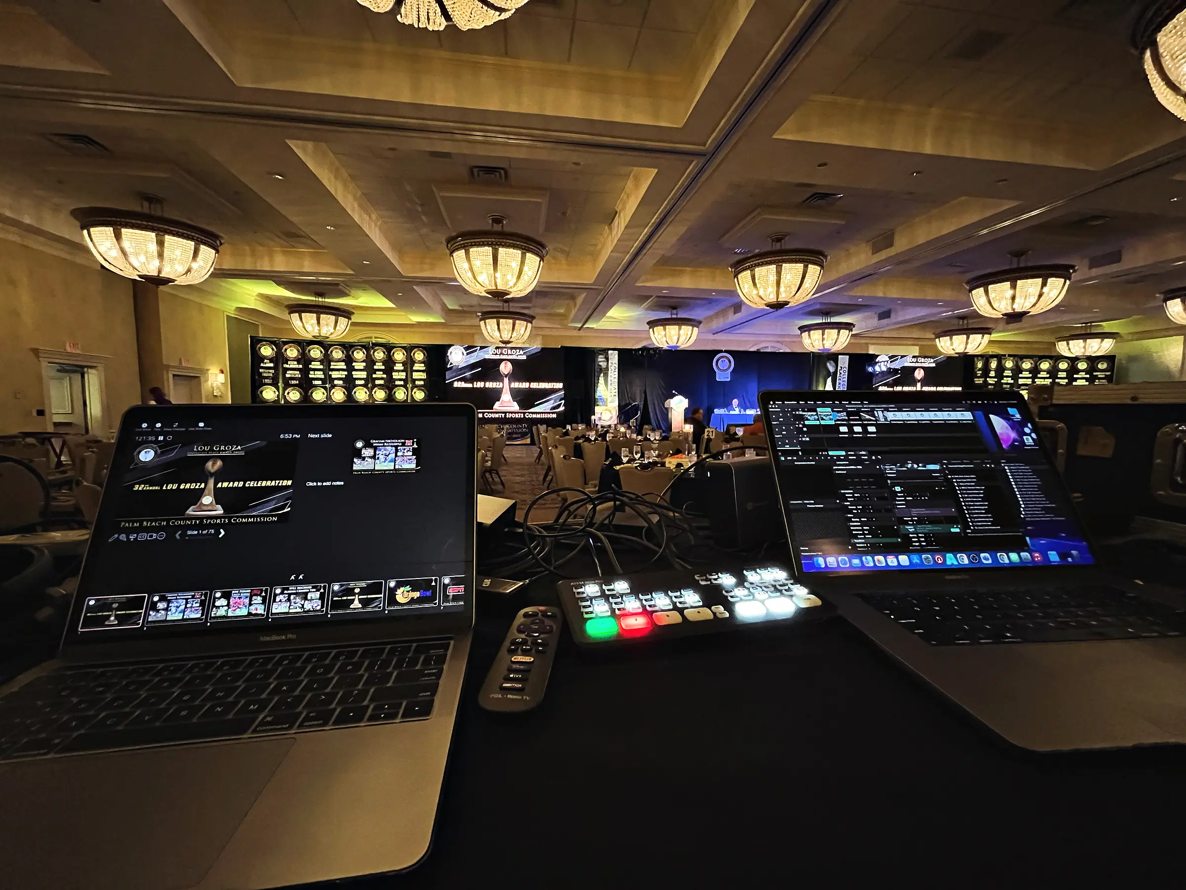 A control position at the back of a ballroom: two laptops open on a draped table with a lit switcher panel between them, and beyond it a wide screen and an awards set under chandeliers.