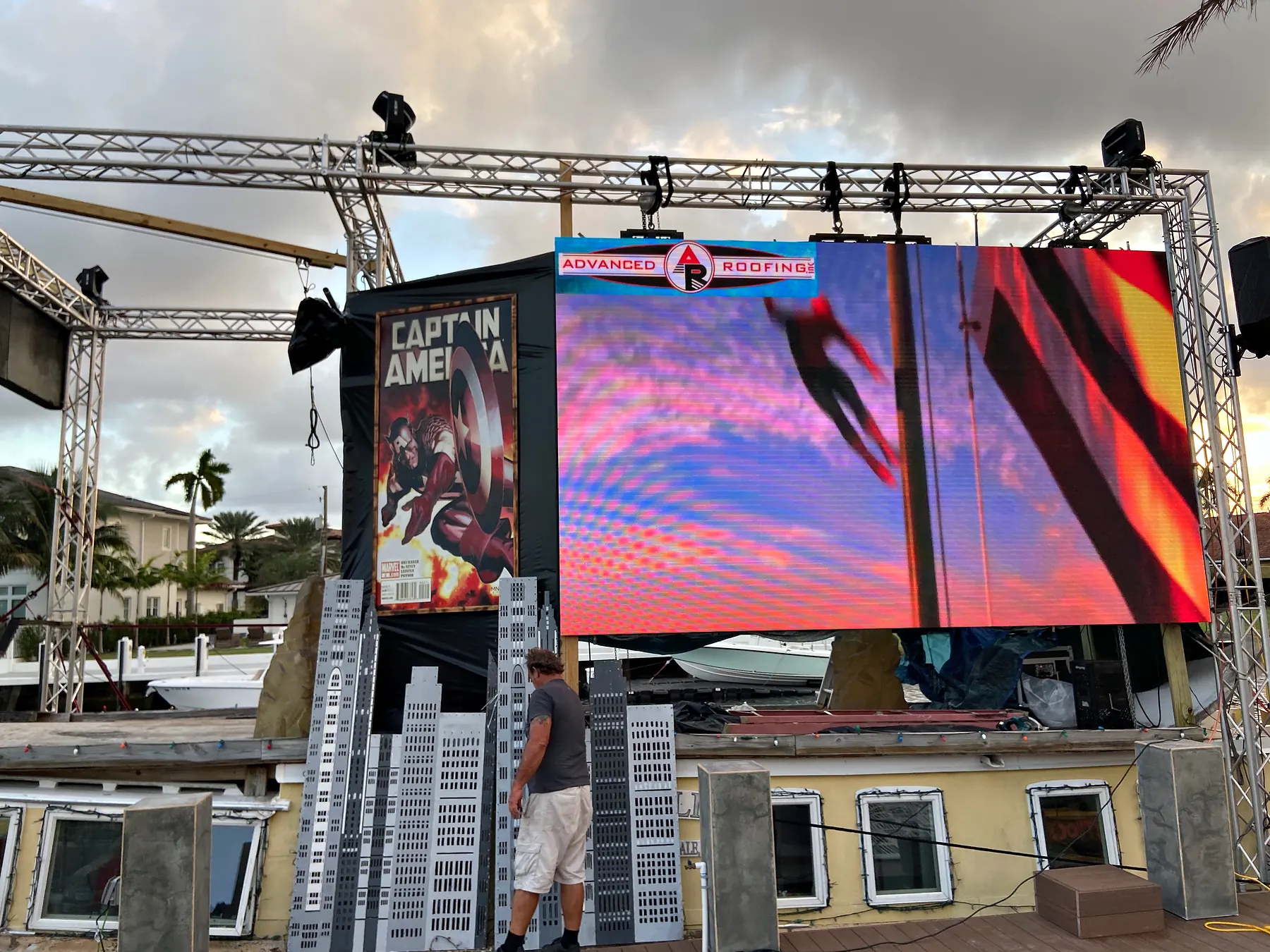 A video wall carried on a ground-supported truss structure built on a barge, loudspeakers hung at the top corners, cut-out skyline scenery standing along the front edge and a tall printed banner at the left, under a heavy evening sky.