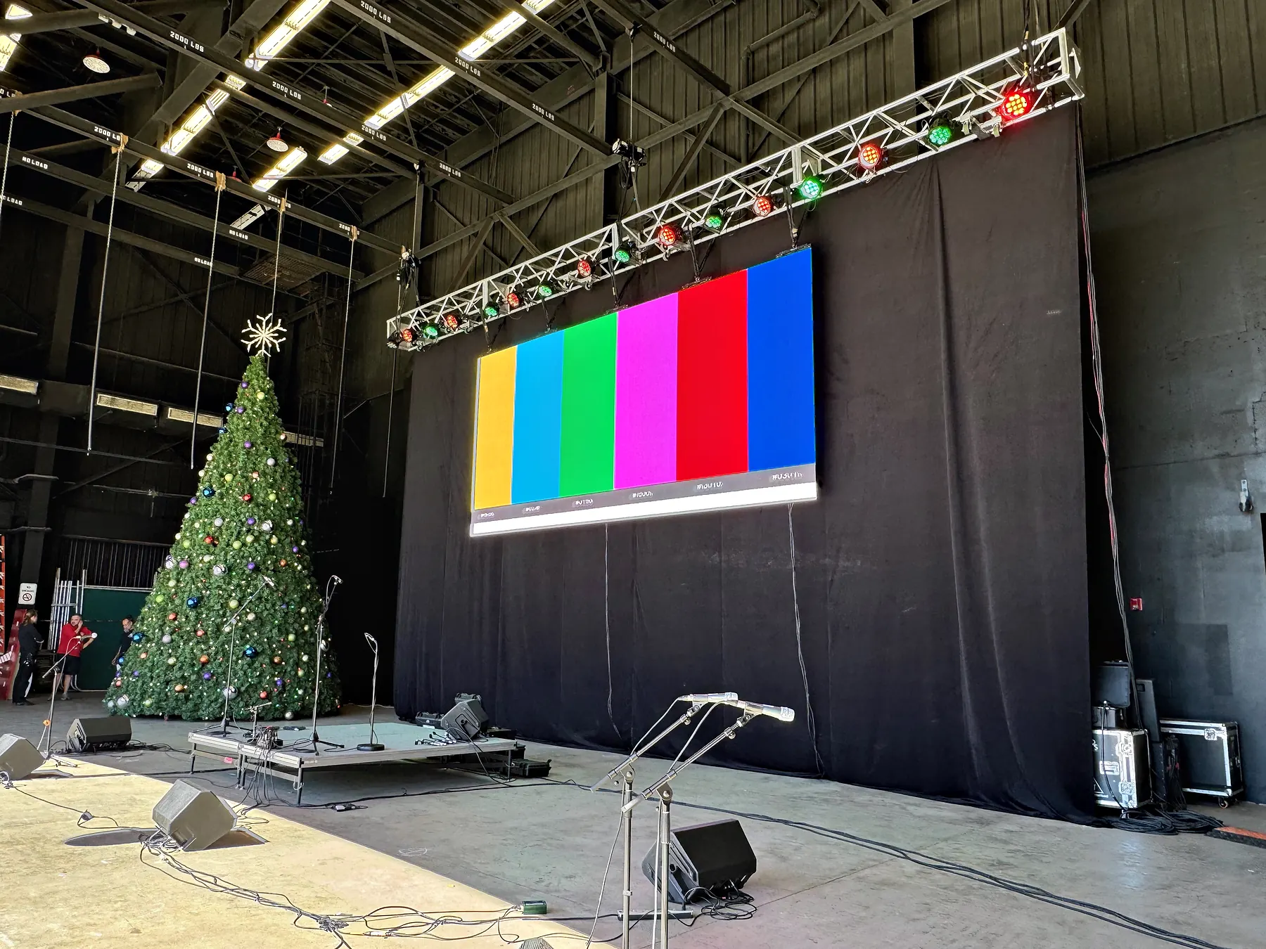 A video wall showing four vertical color bars on a covered stage, a tall decorated tree standing beside it, a lighting truss strung with decorations above and cable and cases across the deck.